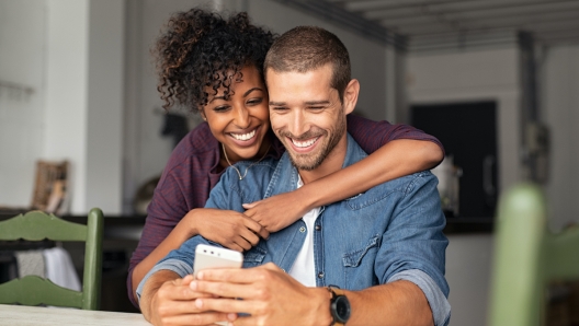 Smiling young couple embracing while looking at smartphone. Multiethnic couple sharing social media on smart phones while sitting at table. Smiling african girl embracing from behind her happy boyfriend while using cellphone and laughing.