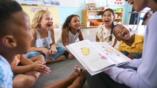 Female Primary Or Elementary School Teacher Reads Story To Multi-Cultural Class Seated In Classroom