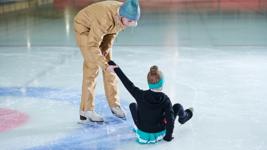 Full length portrait of female coach helping little girl get up during figure skating training in indoor rink, copy space