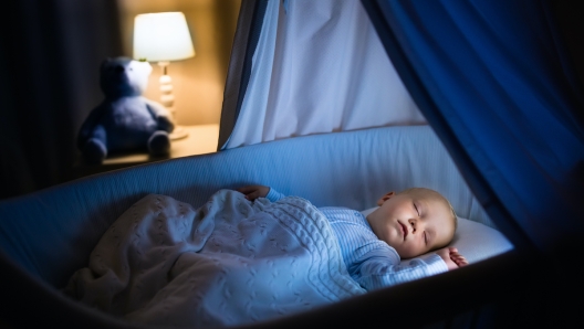 Adorable baby sleeping in blue bassinet with canopy at night. Little boy in pajamas taking a nap in dark room with crib, lamp and toy bear. Bed time for kids. Bedroom and nursery interior.