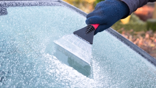 Hand wearing blue glove scratching ice from car window. The ice scraper is an extension of the arm. Part of the windshield is ice free. Only part of the window is visible. Symbol or concept of winter season, cold, frost, weather, sight.