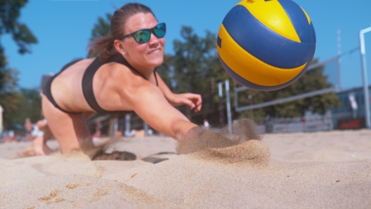 CLOSE UP, DOF: Young female volleyball player dives into the hot sand to save a point during a competitive beach volleyball match. Young Caucasian woman in bikini jumps and hits the ball with one hand