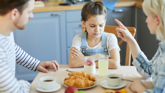 Sad little girl sitting by table while mother forbidding her to have dessert because of bad behavior