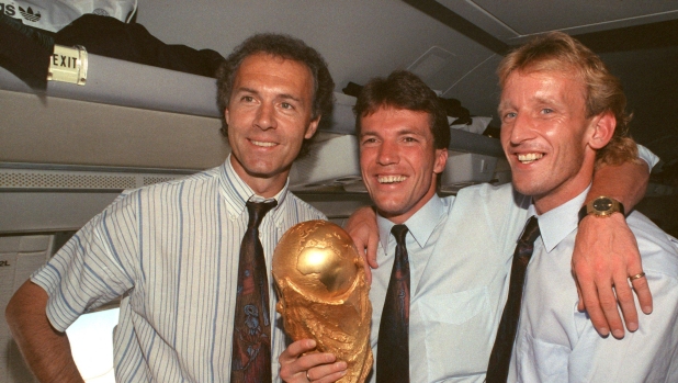 German team leader and head coach Franz Beckenbauer (L) stands with German midfielder and team captain Lothar Matthaeus (C) and German defender Andreas Brehme in the cabin of an airplane and present the World Cup trophy on their way to Germany, 9 July 1990. Germany had won the World Cup final against Argentina in Italy by a score of 1-0 and had won the world championship for the third time since 1954 and 1974. (Photo by Eilmes / DPA / dpa Picture-Alliance via AFP)