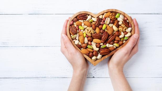Womans hands holding heart shaped bowl with mixed nuts on white table top view. Healthy food and snack.