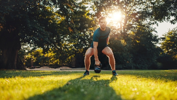 Full length portrait of fitness young man training with kettlebell in the park. Fit caucasian model doing physical workout in the park.