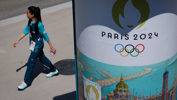 A Paris 2024 volunteer walks behind a Paris 2024 Olympic logo inside the athletes' village for the Paris 2024 Olympics and Paralympics game, in Saint-Denis, north of Paris, on July 18, 2024, ahead of the opening ceremony. The athletes' village for the Paris Olympics and Paralympics officially opened and welcomed its first inhabitants on July 18, 2024. The village to the north of Paris will house nearly 14,500 people, including 9,000 athletes, at its peak. Eight days before the opening ceremony, the first team members to arrive were from Australia and Brazil. (Photo by Geoffroy VAN DER HASSELT / AFP)