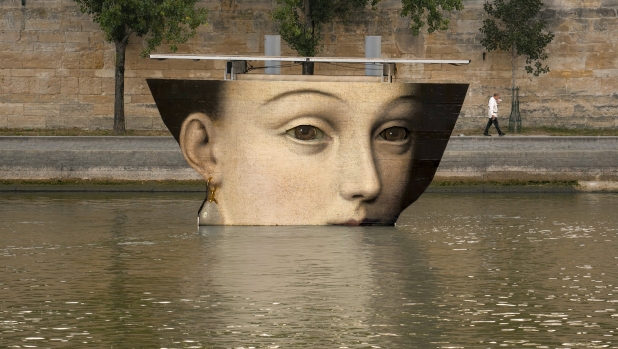 A man wipes his face as he walks past reproductions of artworks decorating the banks of the River Seineat the 2024 Summer Olympics, Tuesday, July 30, 2024, in Paris, France. The men's Olympic triathlon has been postponed over concerns about water quality in Paris' Seine River, where the swimming portion of the race was supposed to take place. (AP Photo/Vadim Ghirda)