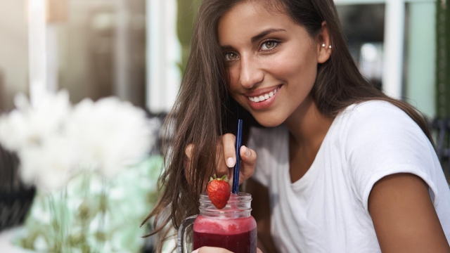 Close-up attractive coquettish tanned european young woman sip strawberry smoothie hold glass drink touch straw smiling tender turn camera sit outdoor street terrace cafe talking.