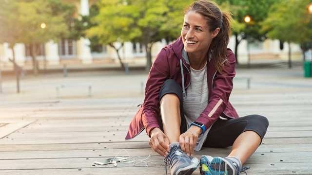Mature fitness woman tie shoelaces on road. Cheerful runner sitting on floor on city streets with mobile and earphones wearing sport shoes. Active latin woman tying shoe lace before running.