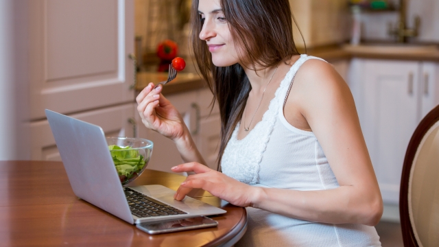 A young beautiful pregnant woman eating a fresh green vegetable salad at the table. Lady having lunch in the kitchen and using the laptop. Healthy nutrition and pregnancy. Healthy lifestyle.