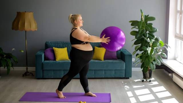 A fat, obese pregnant lady is doing Pilates at home, lifting a fit ball above herself. Home workouts in Interior room with a large window, sofa and yellow-purple pillows. Weight loss in quarantine