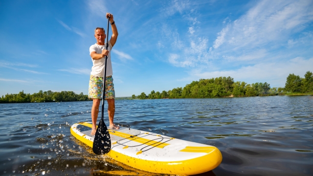 Joyful man is training  SUP board in large river on a sunny morning against a blue sky background . Stand up paddle boarding - awesome active outdoor recreation. Wide angle.