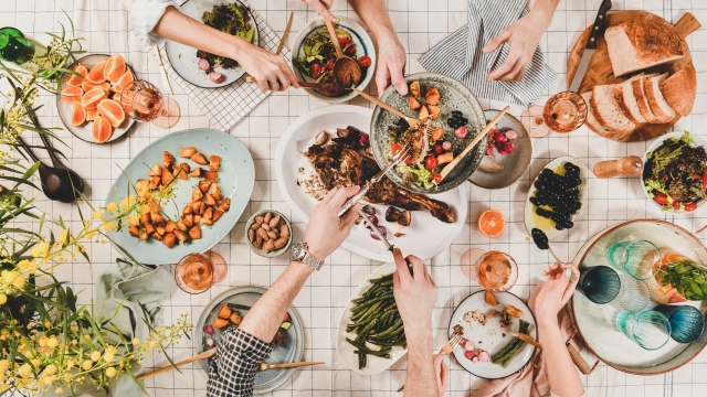 Family or friends gathering dinner. Flat-lay of peoples hands, roasted lamb shoulder, salads, vegetables, rose wine, mimosa branches over white checkered tablecloth, top view. Celebration party dinner