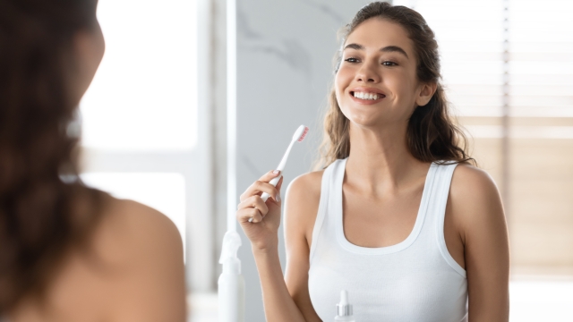 Young Female Cleaning And Brushing Teeth With Toothbrush Standing Near Mirror And Smiling To Reflection In Modern Bathroom Indoor. Toothcare And Oral Hygiene, Toothpaste Advertisement Concept