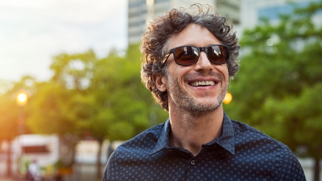 Portrait of smiling man wearing sunglasses and looking away in the city streets. Cheerful mature businessman walking with a big smile on face. Happy man in blue shirt and glasses enjoying the sunset.