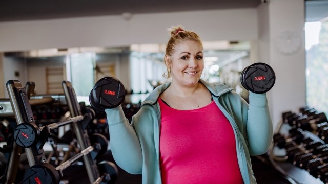 A plus size woman training and lifting dumbbells indoors in gym, looking at camera