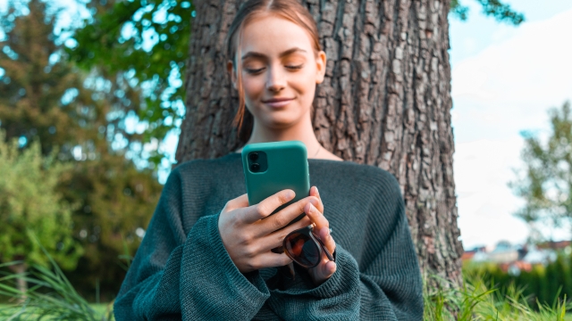Confident smiling teenage woman leaning on tree in the garden, reading messages on her mobile phone, smiling confident and happy. Young women social media lifestyle concept shot.