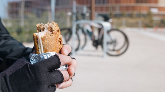 closeup of a young caucasian cyclist man wearing cycling gloves eating a ham and cheese sandwich, wrapped in aluminum foil, next to his bicycle outdoors