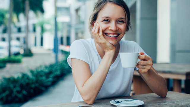 Closeup portrait of smiling young beautiful woman looking at camera, leaning head on hand and drinking coffee at cafe table outdoors with street view in background. Front view.