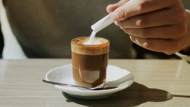 a young man ads some sugar to his machiatto sitting at the table of a sidewalk cafe, in a panoramic format to use as web banner or header
