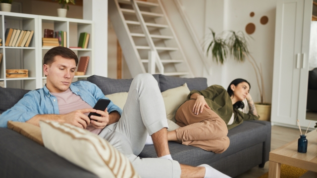 Full length portrait of bored young couple watching TV on sofa at home, focus on man using smartphone in foreground, copy space