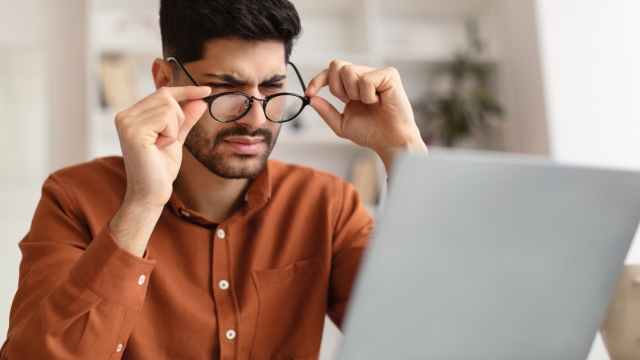 Young male freelancer with bad eyesight using laptop, trying to work from home. Middle Eastern guy holding and taking off his glasses and squinting, looking at laptop screen, having vision troubles
