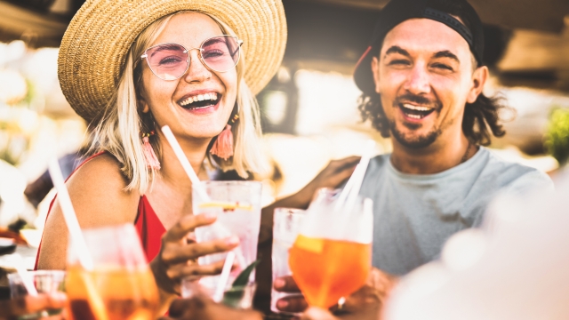 Trendy couple having fun drinking fancy cocktails at beach party - Summer joy and genuine life style concept with young people at festival happy hour - Warm contrast filter with focus on left woman