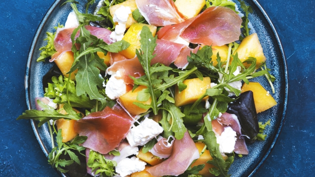 Delicious summer fresh salad with cantaloupe melon, prosciutto, soft cheese and arugula on blue table background, top view, copy space
