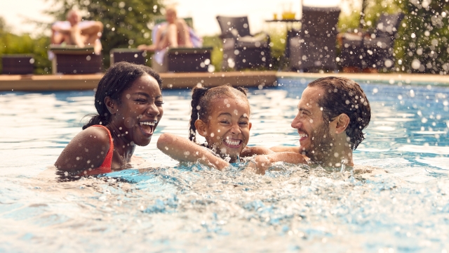 Smiling Mixed Race Family On Summer Holiday Having Fun Splashing In Outdoor Swimming Pool