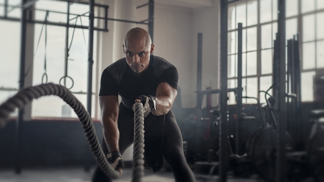 Athlete working out with battle rope at gym. Bald african man training using battle ropes. Fit sportsman doing crossfit exercise in an industrial dark gym.