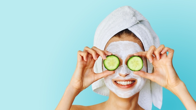 Beautiful young woman with facial mask on her face holding slices of fresh cucumber. Skin care and treatment, spa, natural beauty and cosmetology concept, over blue background with copy space