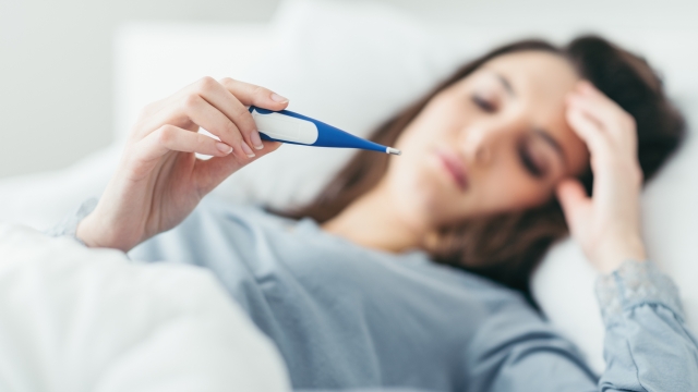 Woman with flu virus lying in bed, she is measuring her temperature with a thermometer and touching her forehead