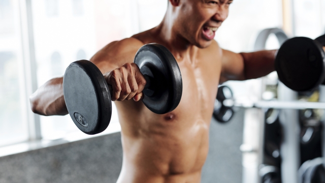 Shirtless fit man shouting when doing side lateral rises with heavy dumbbells