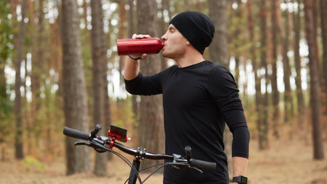 Image of young thirsty athletic sportsman drinking water with closed eyes, having peaceful facial expression, standing in profile, holding his bicycle, being in forest during training. Sport concept.
