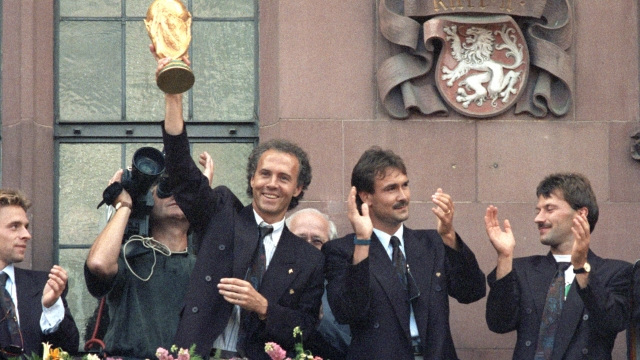 FILED - 09 July 1990, Hesse, Frankfurt\Main: The then team manager Franz Beckenbauer (l) holds up the World Cup trophy he won in Italy on the balcony of the Römer, laughing. He is flanked by (from left): Raimond Aumann and Günther Hermann. Many thousands of people give the players of the German soccer World Cup team an enthusiastic welcome on the Römerberg. The German national soccer team is world champion for the third time. The previous evening, Argentina had been defeated 1:0 in the final in Rome. Franz Beckenbauer is dead. The German soccer legend died on Sunday at the age of 78, his family told the German Press Agency on Monday. Photo: Kai-Uwe Wärner/dpa (Photo by Kai-Uwe Wärner / DPA / dpa Picture-Alliance via AFP)