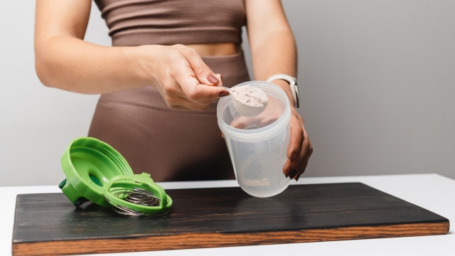 Athletic woman in sportswear with measuring spoon in her hand puts portion of whey protein powder into a shaker on a table, making protein drink cocktail.