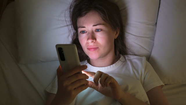 Portrait of a young woman who lies on the bed at night and types messages on cell phone.
