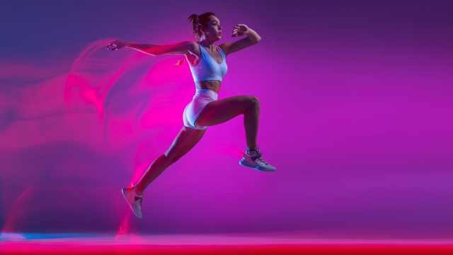 Professional female athlete, runner training isolated on blue studio background in mixed pink neon light. Sportive girl in white sportswear practicing in run or jogging. Healthy lifestyle concept.