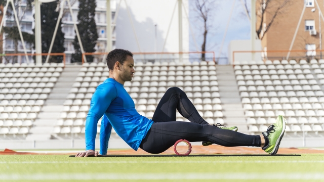 Stock photo of african american male athlete sitting on the track stretching his legs with foam roller.