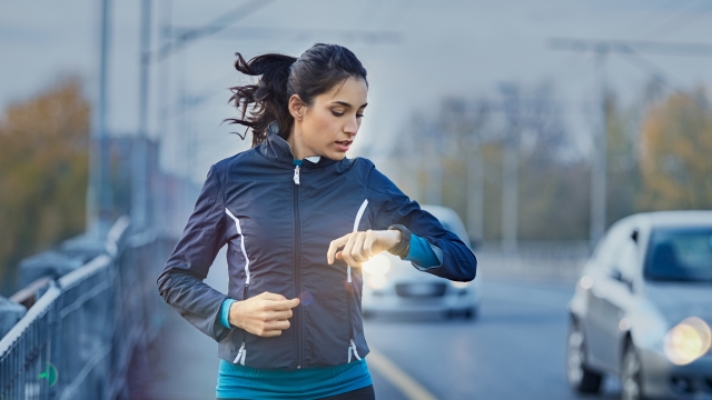 Young fitness woman runner checking time from smart watch. Young woman checking heart rate while jogging on street at dusk. Young woman looking on smartwatch her heartbeat while running in city.