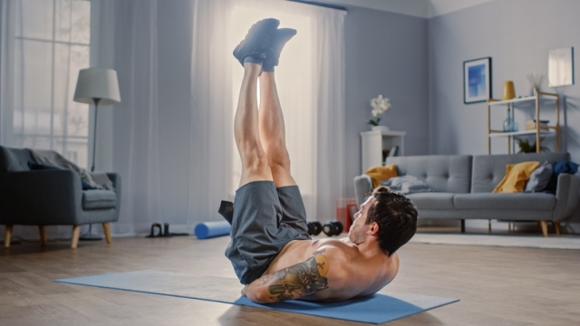 Muscular Athletic Shirtless Fit Man in Grey Shorts Injures His Back After Lifting Dumbbells at Home in His Spacious and Sunny Living Room with Minimalistic Interior.