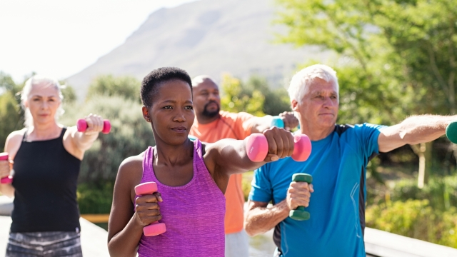 Happy senior and mature couples exercising with dumbbells. Healthy multiethnic people exercising using dumbbells outdoor. African couple and senior friends in sportswear stretching arms holding colorful dumbbells at park.