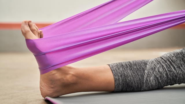 Crop unrecognizable female athlete sitting on yoga mat and stretching leg with elastic band placed around foot in modern gym