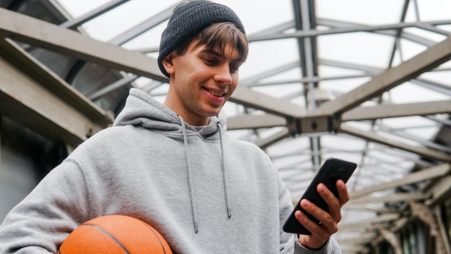 Young man basketball player with headphones holding ball using smartphone after training. Urban background.