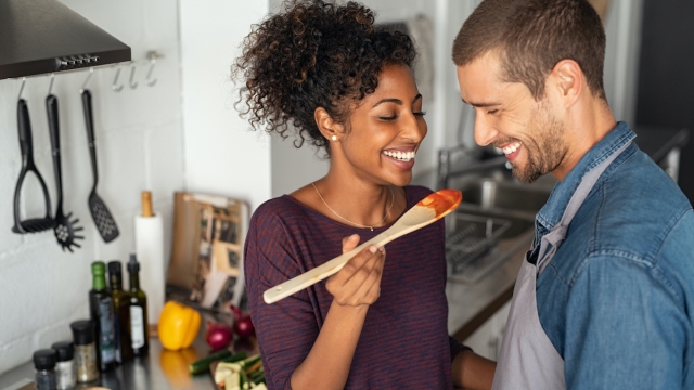 Young couple tasting tomato sauce while cooking in the kitchen. Cheerful man and smiling woman holding spatula in hand ready to taste red sauce. Multiethnic couple cooking together at home.
