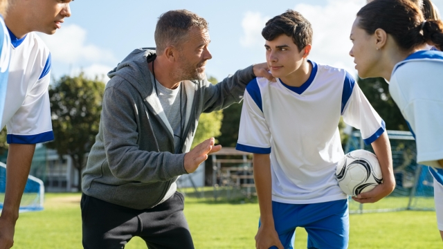 Mature coach teaching strategy to high school team on football field. Young soccer players standing together united and listening coach motivational speech. Coach giving team advise before school match.