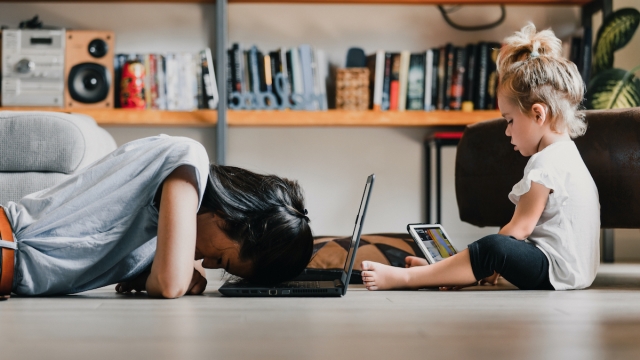 Young woman, presumably a mother, is lying on the floor with her head on a laptop, appearing exhausted, while her daughter is sitting next to her playing on a tablet