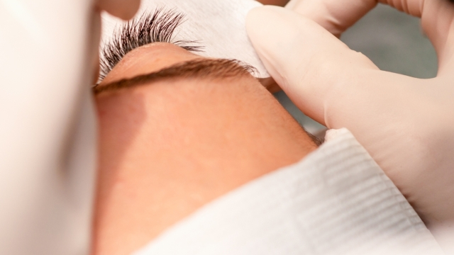 The hands of the cosmetologist are gluing white tape under the eye of the young caucasian woman during the eyelash extension procedure, closeup