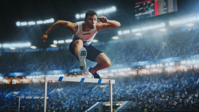 Professional Sport Performance: Talented Male Hurdler Jumping Over Obstacle, Racing Against Time and Setting a New Sprint Record in Front of a Stadium with Cheering Fans During National Competition